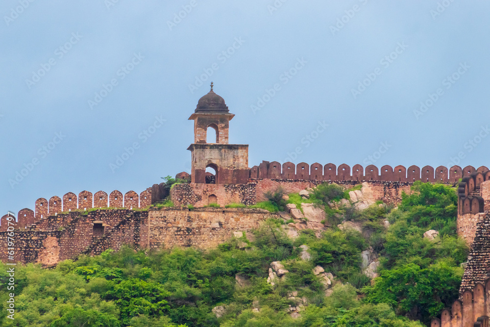 Dome-shaped watch tower on the fortified wall of an ancient Indian fort on the top of the mountain covered with lush green forest amid the clear blue sky. 