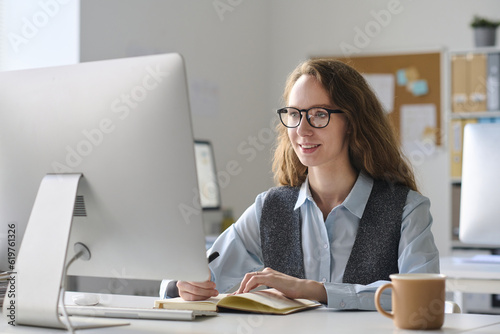 Young businesswoman in eyeglasses working on computer at her workplace and making notes in notepad