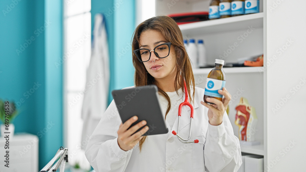Young beautiful hispanic woman doctor using touchpad holding medication bottle thinking at clinic