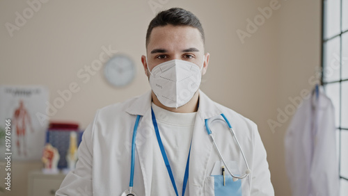 Young hispanic man doctor standing with serious expression wearing medical mask at clinic