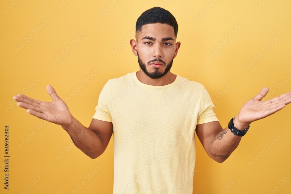 Young hispanic man standing over yellow background clueless and confused expression with arms and hands raised. doubt concept. Young hispanic man standing over yellow background clueless and confused expression with arms and hands raised. doubt concept.