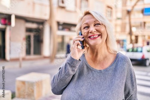 Middle age blonde woman smiling confident talking on the smartphone at street