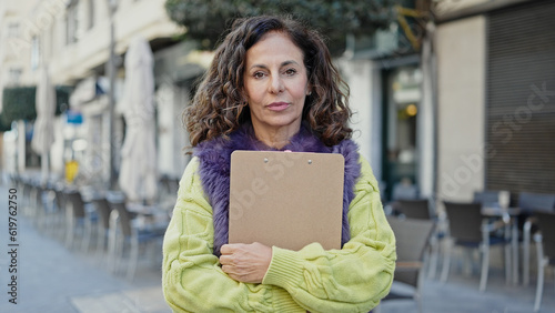 Middle age hispanic woman standing with serious expression holding clipboard at coffee shop terrace