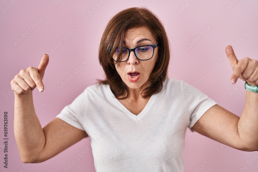Middle age hispanic woman standing over pink background pointing down with fingers showing advertisement, surprised face and open mouth Middle age hispanic woman standing over pink background pointing down with fingers showing advertisement, surprised face and open mouth