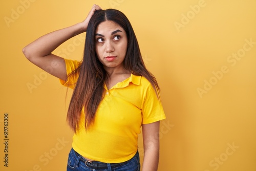 Young arab woman standing over yellow background confuse and wondering about question. uncertain with doubt, thinking with hand on head. pensive concept.