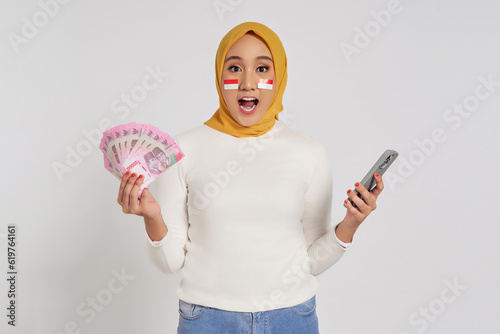 Surprised young Asian Muslim woman wearing a hijab holding money and mobile phone isolated over white background. Celebrate Indonesian independence day on 17 August