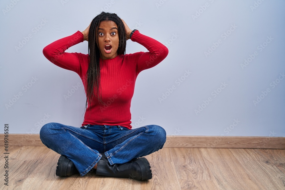 Young african american with braids sitting on the floor at home crazy and scared with hands on head, afraid and surprised of shock with open mouth Young african american with braids sitting on the floor at home crazy and scared with hands on head, afraid and surprised of shock with open mouth
