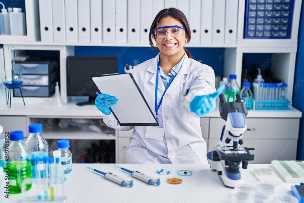 Hispanic young woman working at scientist laboratory smiling cheerful offering palm hand giving assistance and acceptance. Hispanic young woman working at scientist laboratory smiling cheerful offering palm hand giving assistance and acceptance.