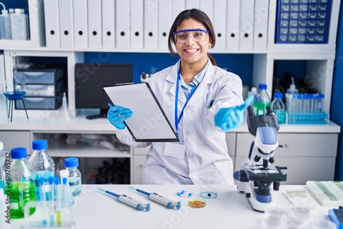 Hispanic young woman working at scientist laboratory smiling cheerful offering palm hand giving assistance and acceptance.