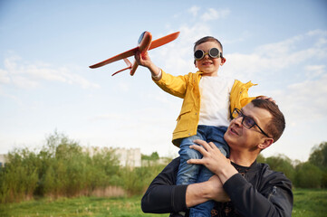 Boy is sitting on man's shoulders. Father with his little son playing with toy plane on the field