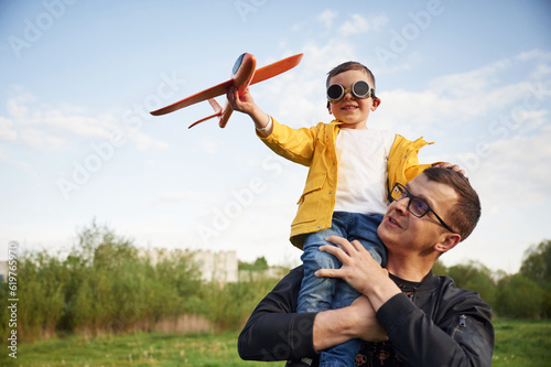 Boy is sitting on man's shoulders. Father with his little son playing with toy plane on the field photo