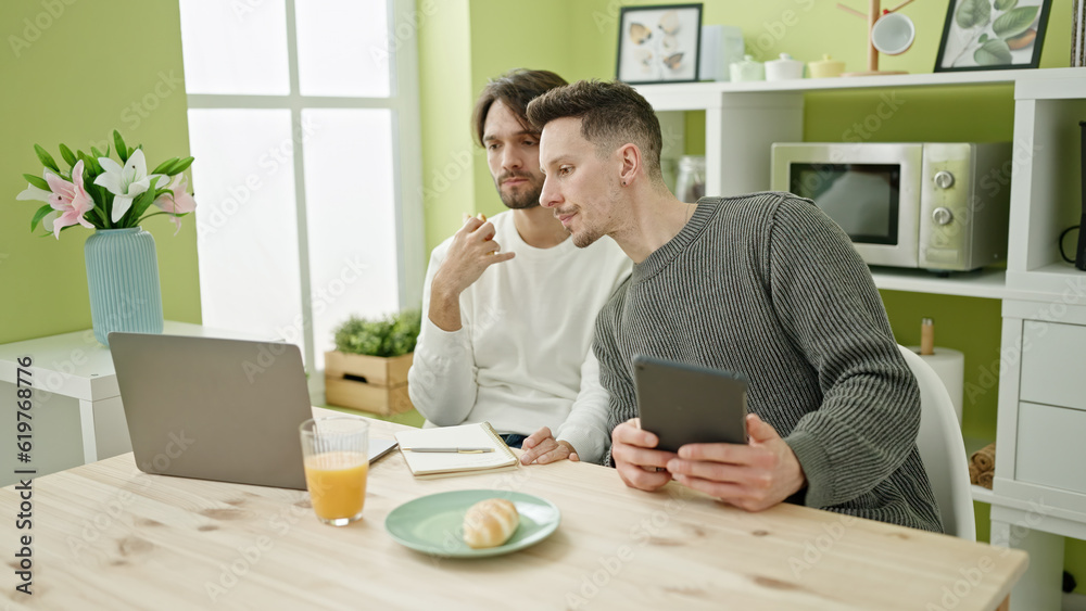 Two men couple having breakfast using touchpad and laptop at dinning room