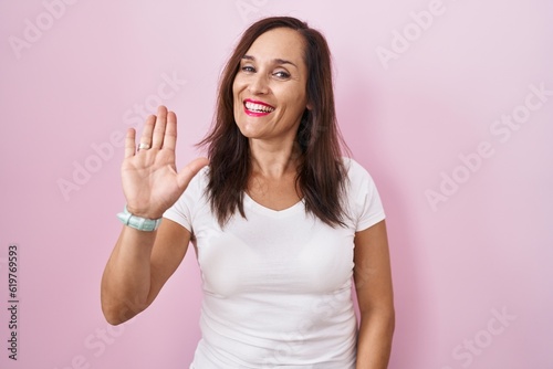 Middle age brunette woman standing over pink background waiving saying hello happy and smiling  friendly welcome gesture