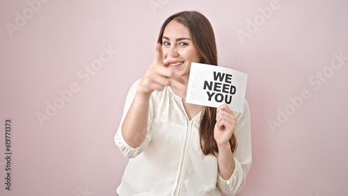 Young beautiful hispanic woman doing coming gesture with hand holding we need you message paper over isolated pink background