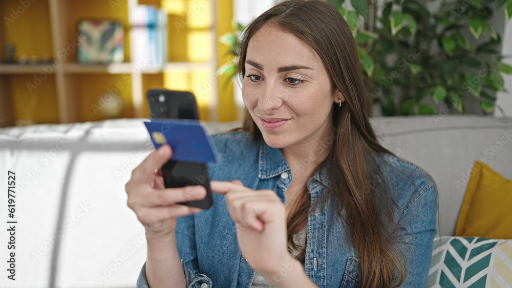 Young beautiful hispanic woman shopping with smartphone and credit card sitting on sofa at home