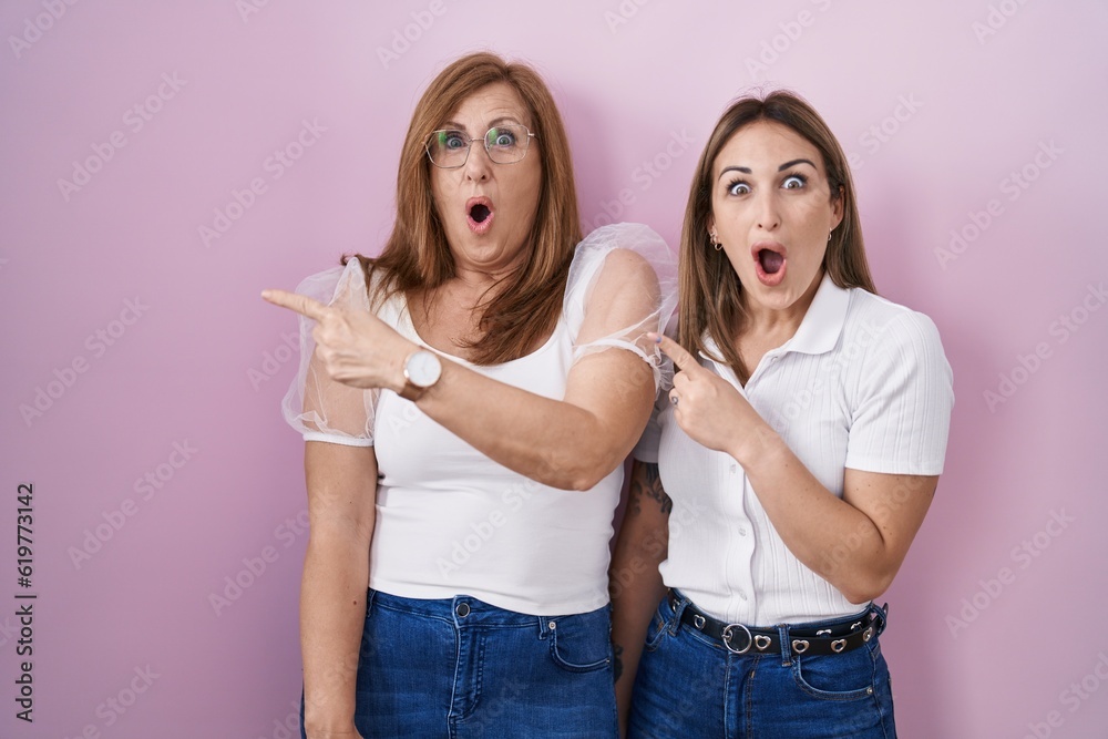 Hispanic mother and daughter wearing casual white t shirt over pink background surprised pointing with finger to the side, open mouth amazed expression. Hispanic mother and daughter wearing casual white t shirt over pink background surprised pointing with finger to the side, open mouth amazed expression.
