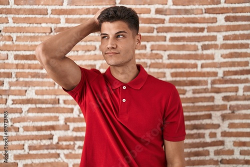 Young hispanic man standing over bricks wall confuse and wonder about question. uncertain with doubt, thinking with hand on head. pensive concept.