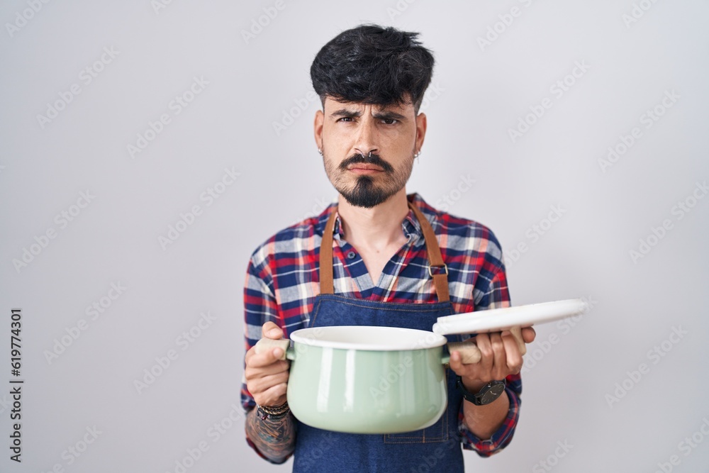 Young hispanic man with beard wearing apron holding cooking pot skeptic and nervous, frowning upset because of problem. negative person.