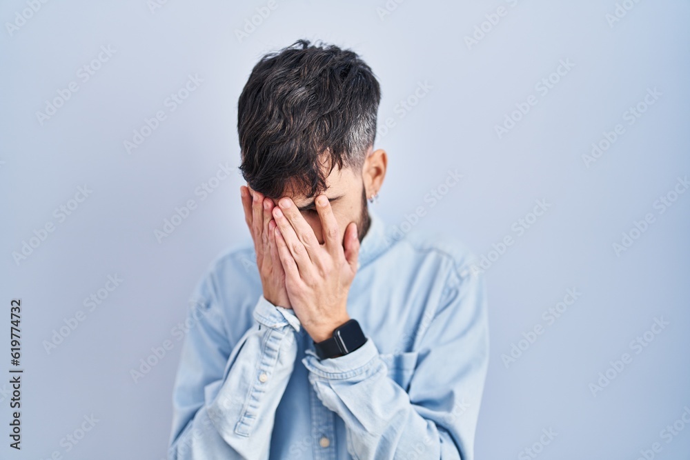 Young hispanic man with beard standing over blue background with sad expression covering face with hands while crying. depression concept. Young hispanic man with beard standing over blue background with sad expression covering face with hands while crying. depression concept.