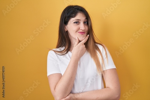 Young brunette woman standing over yellow background looking confident at the camera smiling with crossed arms and hand raised on chin. thinking positive.