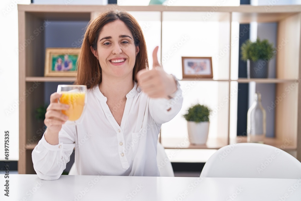 Brunette woman drinking glass of orange juice approving doing positive gesture with hand, thumbs up smiling and happy for success. winner gesture. Brunette woman drinking glass of orange juice approving doing positive gesture with hand, thumbs up smiling and happy for success. winner gesture.
