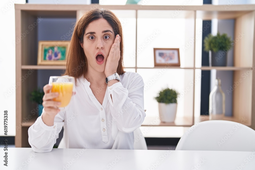 Brunette woman drinking glass of orange juice afraid and shocked, surprise and amazed expression with hands on face Brunette woman drinking glass of orange juice afraid and shocked, surprise and amazed expression with hands on face