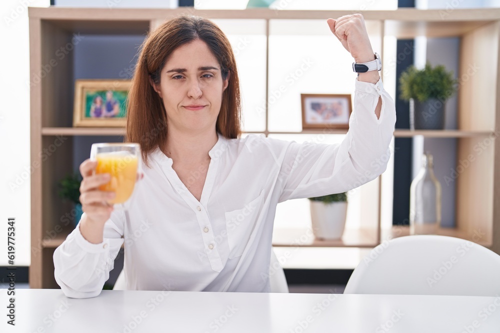 Brunette woman drinking glass of orange juice strong person showing arm muscle, confident and proud of power Brunette woman drinking glass of orange juice strong person showing arm muscle, confident and proud of power