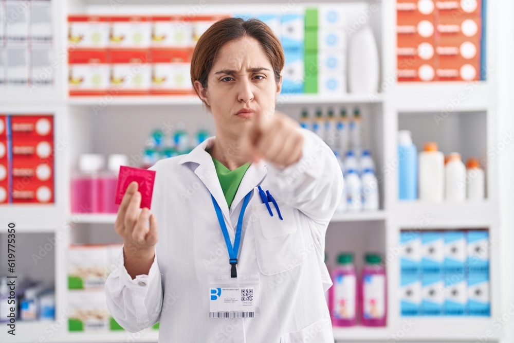 Brunette woman working at pharmacy drugstore holding condom pointing with finger to the camera and to you, confident gesture looking serious