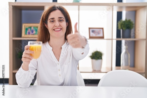 Brunette woman drinking glass of orange juice approving doing positive gesture with hand, thumbs up smiling and happy for success. winner gesture.