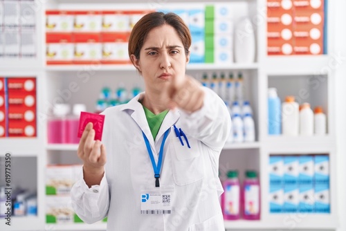 Brunette woman working at pharmacy drugstore holding condom pointing with finger to the camera and to you  confident gesture looking serious