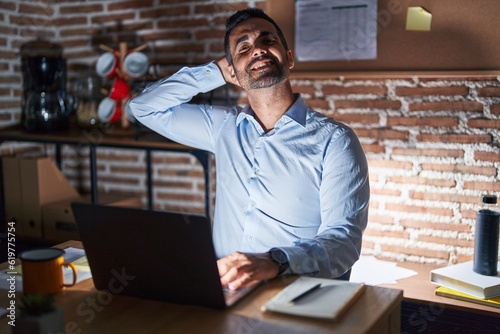 Hispanic man with beard working at the office at night smiling confident touching hair with hand up gesture, posing attractive and fashionable