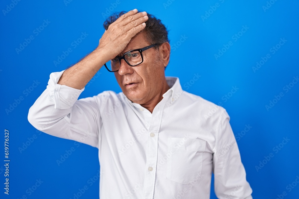 Middle age hispanic man standing over blue background surprised with hand on head for mistake, remember error. forgot, bad memory concept. Middle age hispanic man standing over blue background surprised with hand on head for mistake, remember error. forgot, bad memory concept.