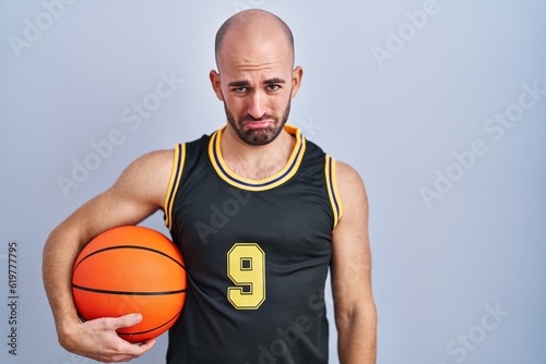 Young bald man with beard wearing basketball uniform holding ball depressed and worry for distress, crying angry and afraid. sad expression.