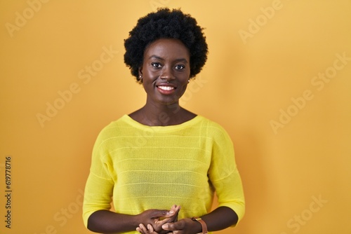 African young woman standing over yellow studio with hands together and crossed fingers smiling relaxed and cheerful. success and optimistic © Krakenimages.com