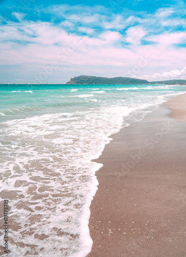 Poetto beach in Cagliari, Sardinia is a beautiful destination with small blue waves and white foam contrasting with the sand. The promontory of Sella del Diavolo and colorful sails are visible.