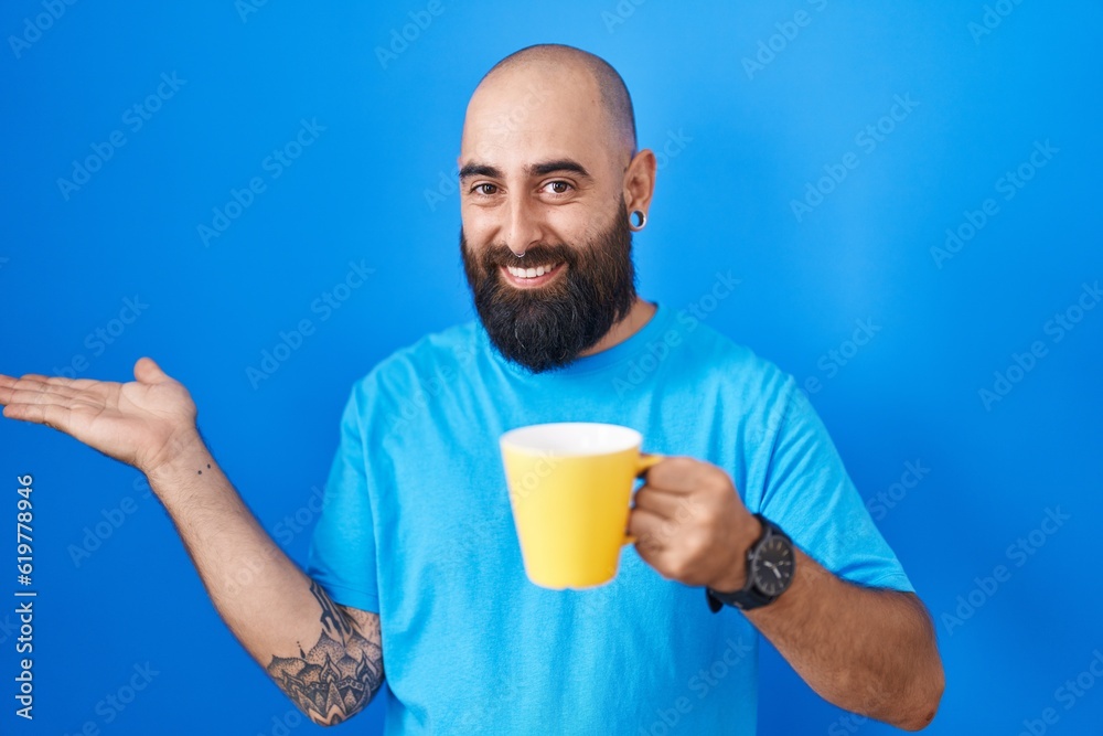 Young hispanic man with beard and tattoos drinking a cup of coffee smiling cheerful presenting and pointing with palm of hand looking at the camera. Young hispanic man with beard and tattoos drinking a cup of coffee smiling cheerful presenting and pointing with palm of hand looking at the camera.