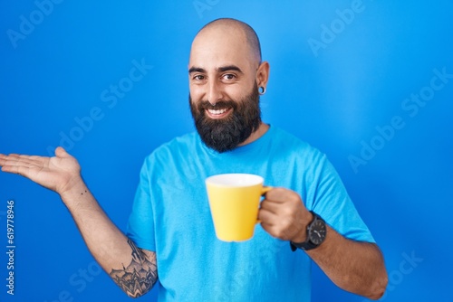 Young hispanic man with beard and tattoos drinking a cup of coffee smiling cheerful presenting and pointing with palm of hand looking at the camera. © Krakenimages.com