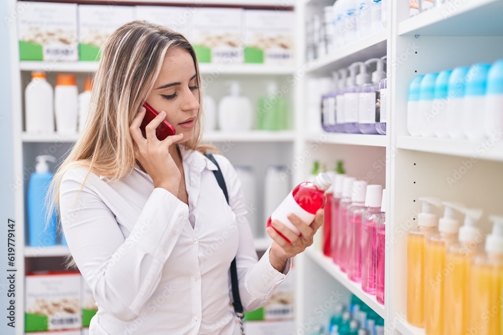 Young blonde woman customer talking on smartphone holding medicine bottle at pharmacy