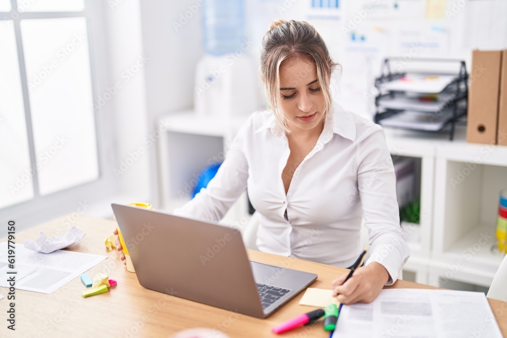 Young blonde woman business worker using laptop writing on reminder paper at office