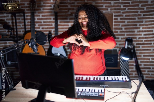 Plus size hispanic woman playing piano at music studio smiling in love doing heart symbol shape with hands. romantic concept.