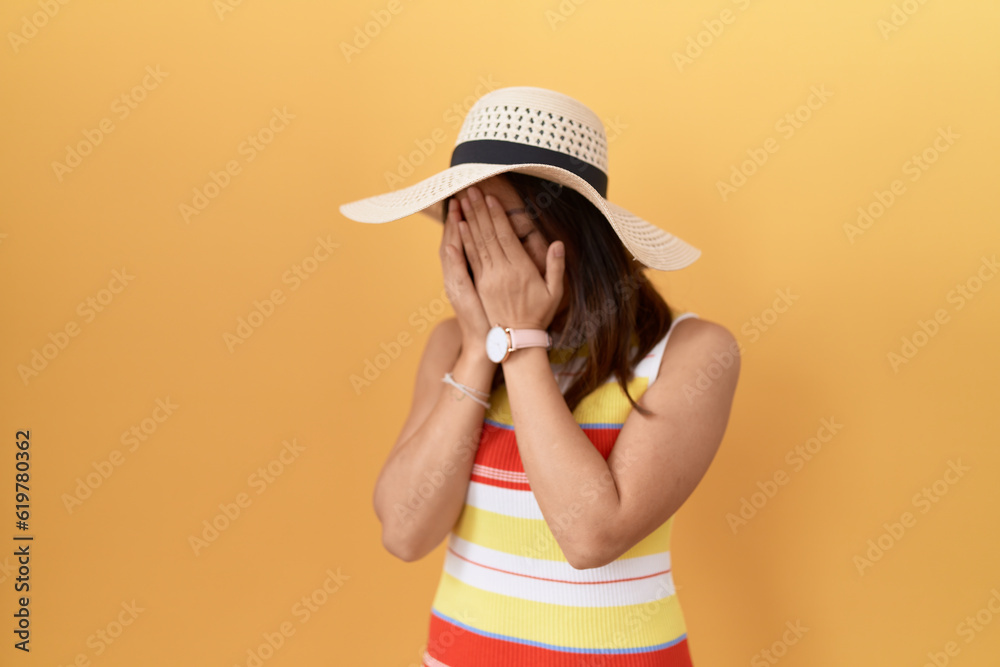 Middle age chinese woman wearing summer hat over yellow background with sad expression covering face with hands while crying. depression concept. Middle age chinese woman wearing summer hat over yellow background with sad expression covering face with hands while crying. depression concept.
