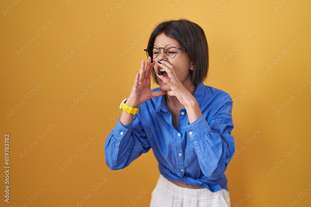 Young girl standing over yellow background shouting angry out loud with hands over mouth Young girl standing over yellow background shouting angry out loud with hands over mouth