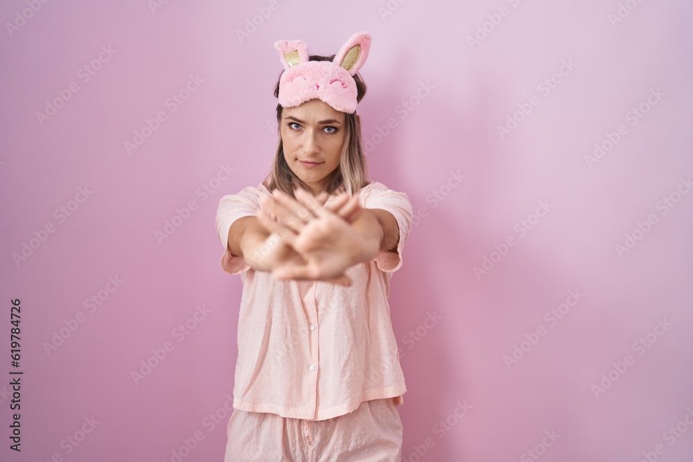 Fototapeta premium Blonde caucasian woman wearing sleep mask and pajama rejection expression crossing arms and palms doing negative sign, angry face
