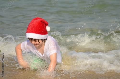 Fototapeta Naklejka Na Ścianę i Meble -  Cute boy in santa hat on the beach. The concept of vacation at sea, family holidays in December. Child on vacation.