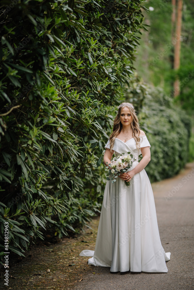 A beautiful bride with a long dress stands in nature in the green with a large bouquet. Wedding portrait of a young bride. Wedding photography.