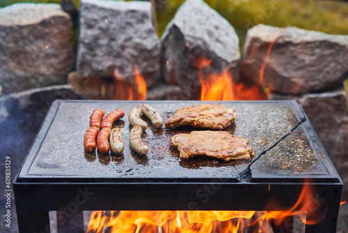 Old fashion barbeque on granite stone slab over the open campfire heated by flames from wood, with two kind of sausages, normal and bvarian style white sausages and two pork collar marinated steaks. 