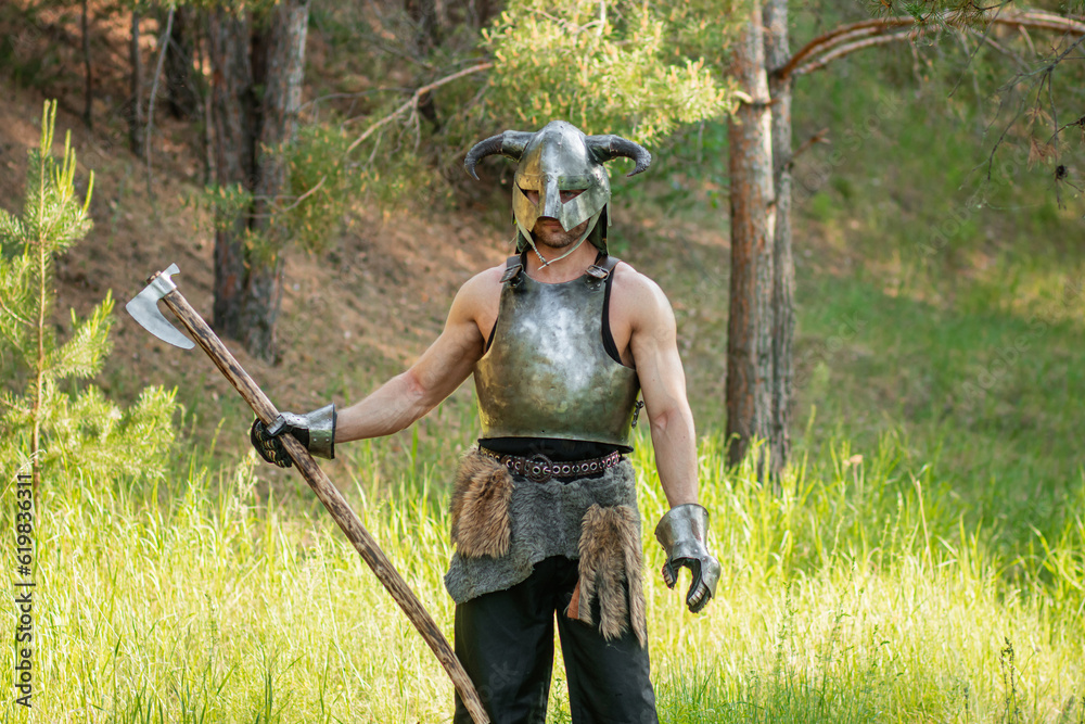 Fototapeta premium Portrait of a warrior in a horned helmet, a steel breastplate with a two-handed ax in his hands, posing against the backdrop of the forest.
