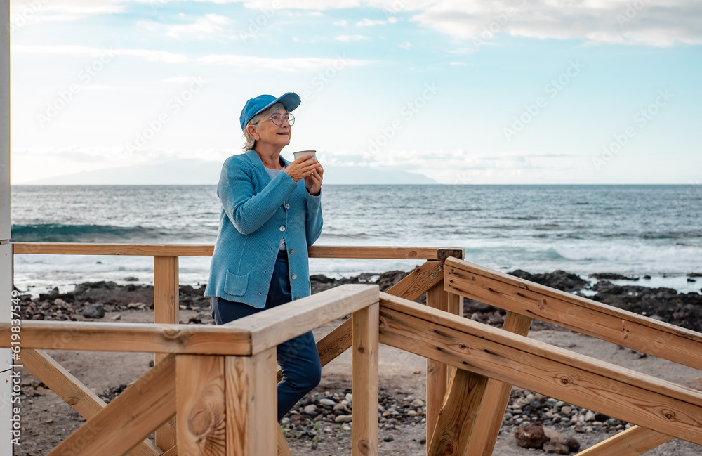 Smiling senior woman in blue standing outdoors at the beach in sunset light holding a coffee cup looking away. Happy pensioner woman enjoying free lifestyle in retirement Smiling senior woman in blue standing outdoors at the beach in sunset light holding a coffee cup looking away. Happy pensioner woman enjoying free lifestyle in retirement