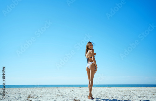 Perfect beach getaway. Gorgeous suntan bikini model turning around to camera, relaxing under warm sun on the deserted island in front sea water against blue sky.