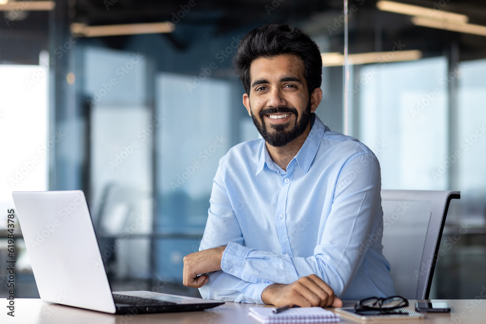 Portrait of happy and successful businessman, indian man smiling and looking at camera, satisfied with achievement results man working inside office building using laptop at work Portrait of happy and successful businessman, indian man smiling and looking at camera, satisfied with achievement results man working inside office building using laptop at work
